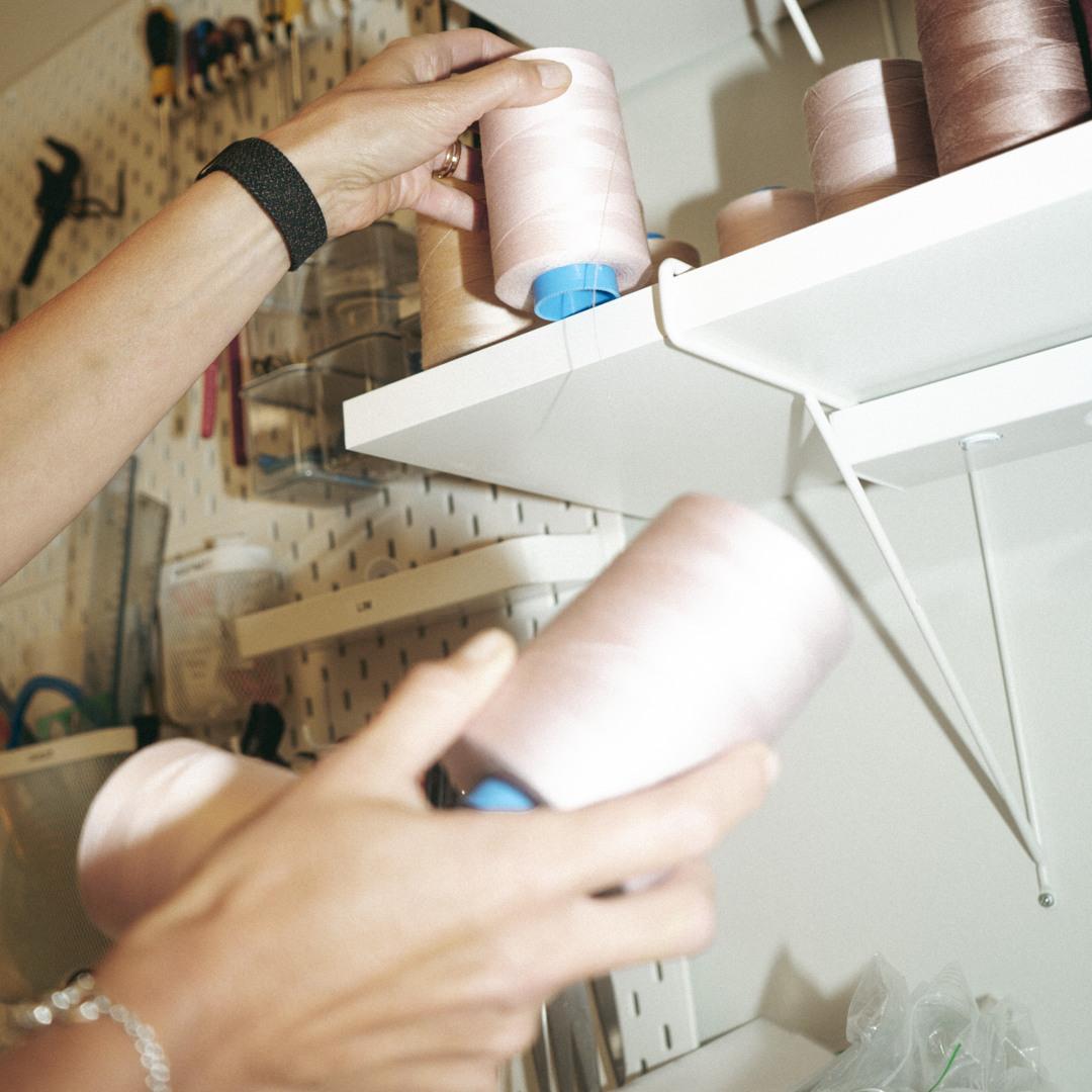 Hands placing pink thread spools on a white shelf with a pegboard wall in the background holding various tools.