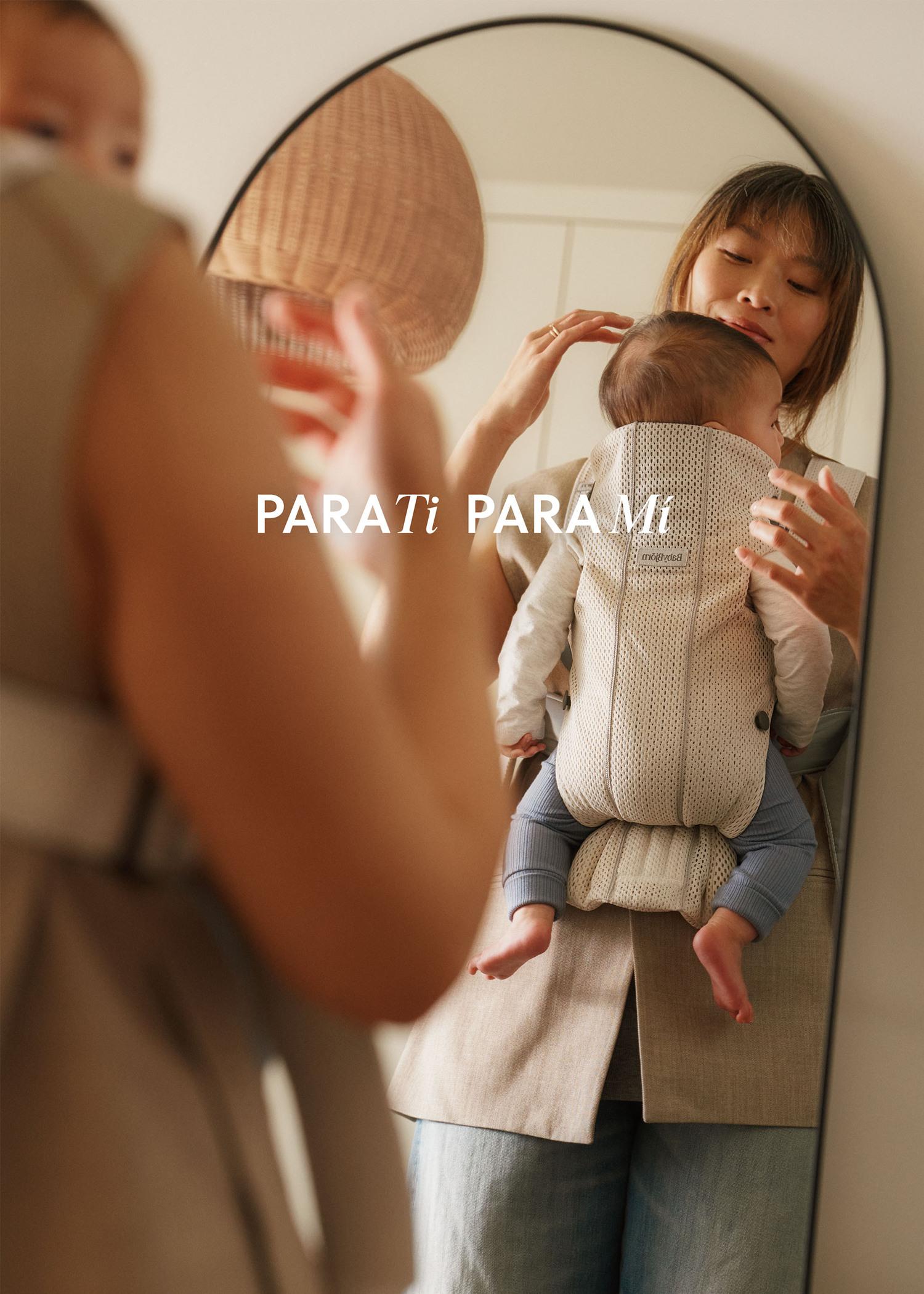 Woman holding a baby in a carrier, looking in a mirror. Text says "PARA Ti PARA Mi." Both are dressed in neutral tones.
