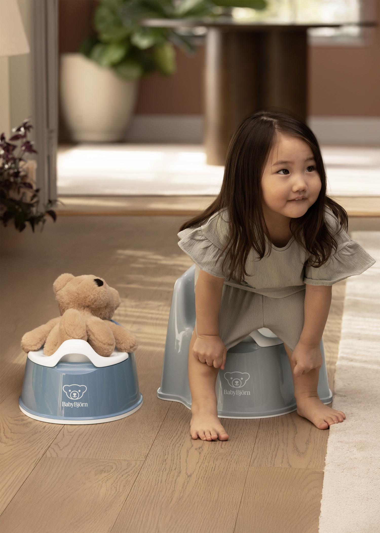 A young girl sits on a gray potty chair, smiling, with a teddy bear on a similar chair beside her, in a cozy room.