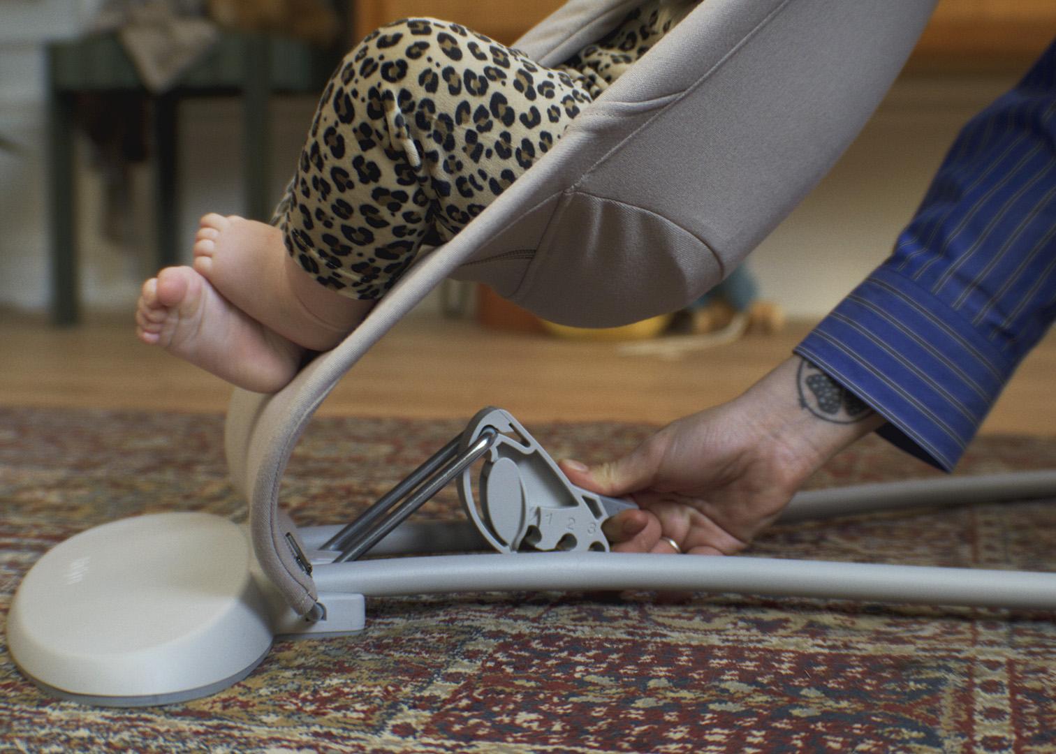 A person adjusts a baby bouncer with a child in leopard print leggings inside. The scene is set on a patterned rug.