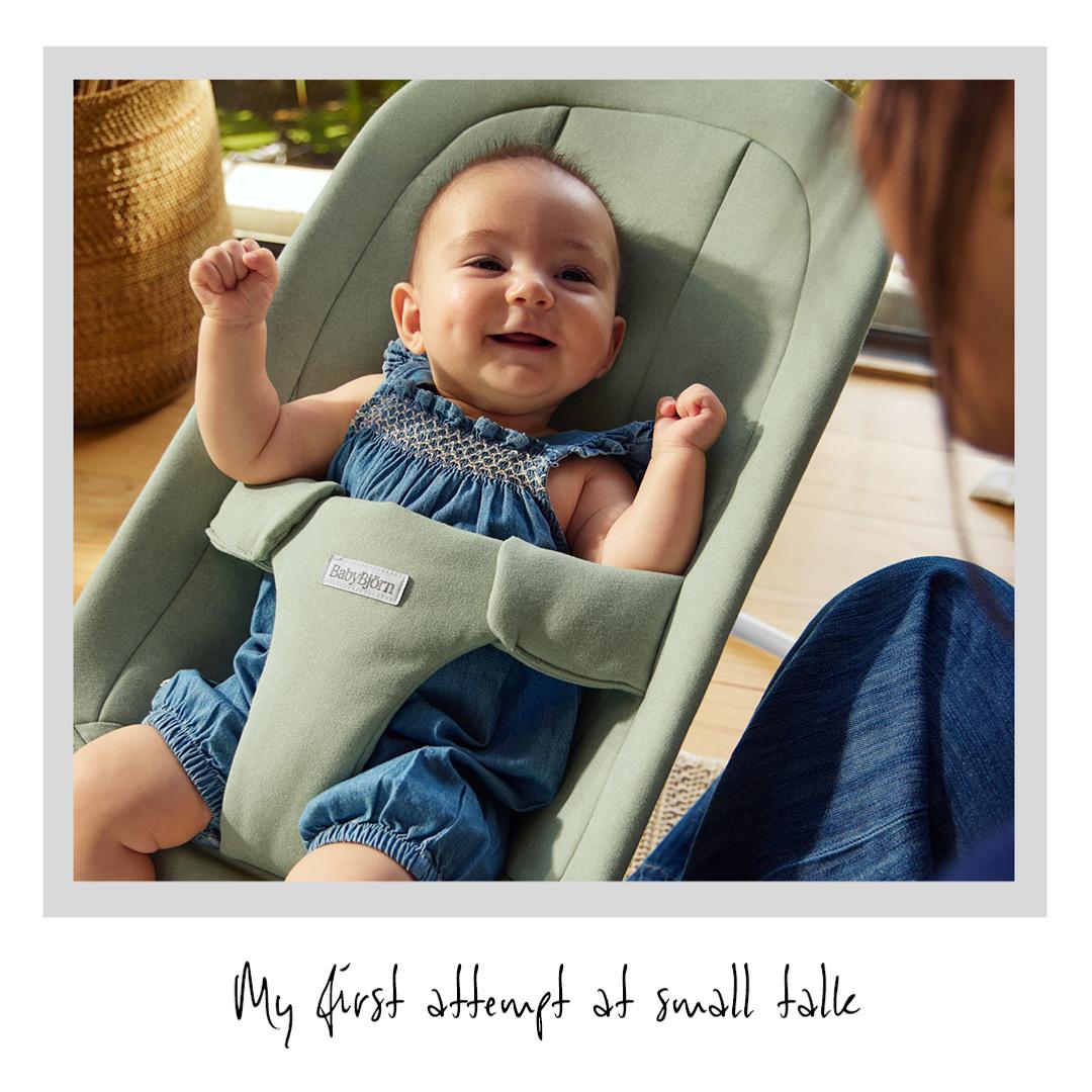 Smiling baby in a green bouncer chair, wearing a blue romper, with an adult nearby. Handwritten text: "My first attempt at small talk."