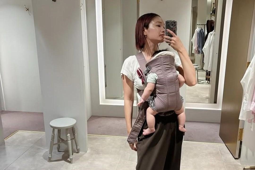 Woman taking a mirror selfie while carrying a baby in a front-facing carrier in a fitting room with clothes and a stool in the background.