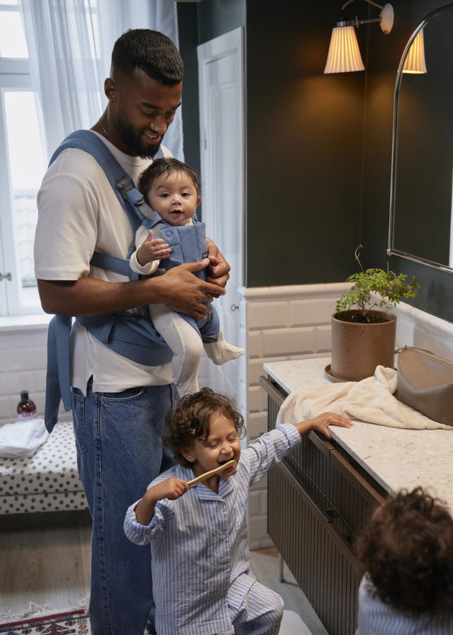 Father holds a baby in a blue baby carrier while a toddler brushes their teeth at a bathroom sink.