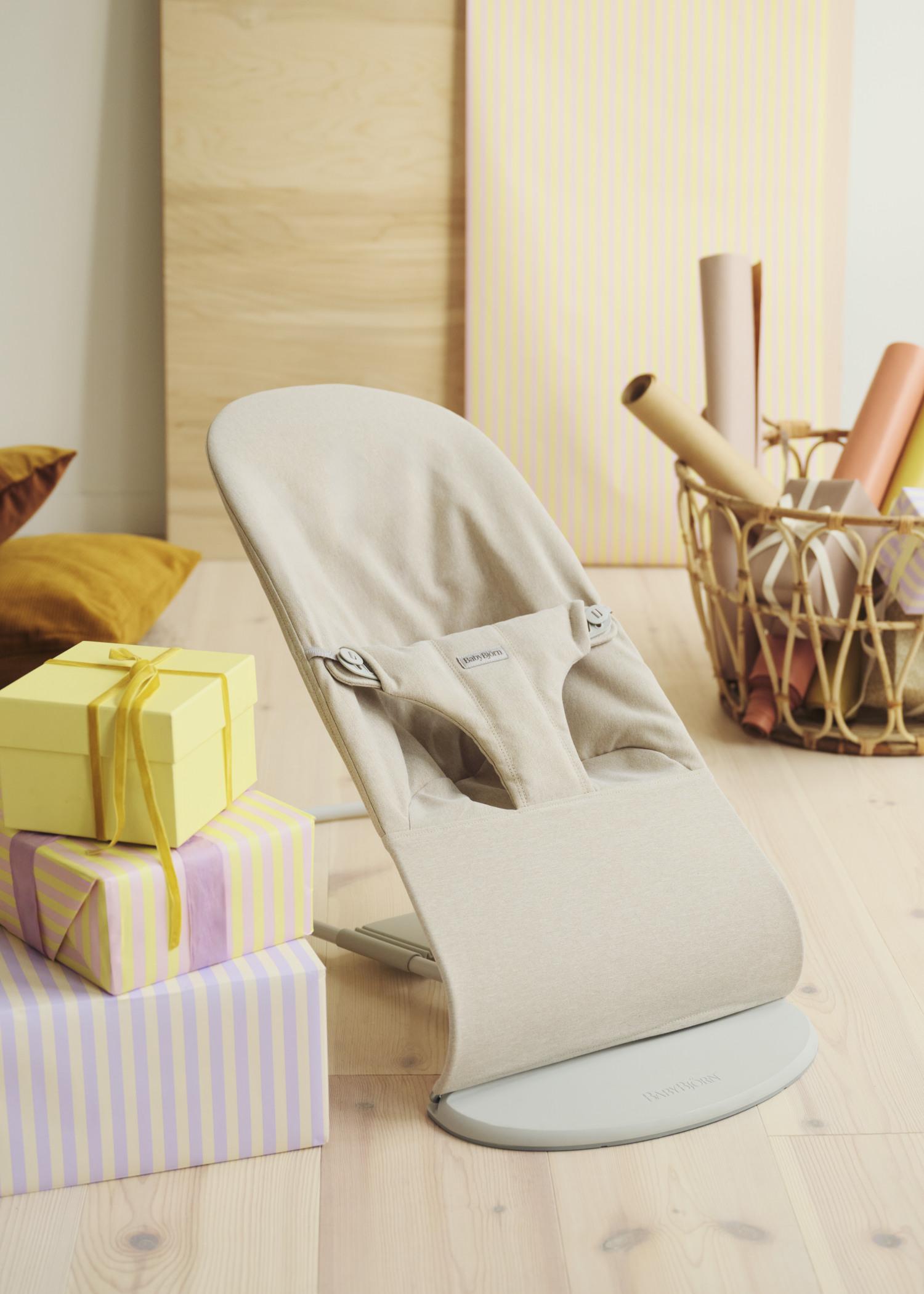 A beige baby bouncer on a light wooden floor, next to wrapped gifts in pastel colors and a basket with rolled papers.