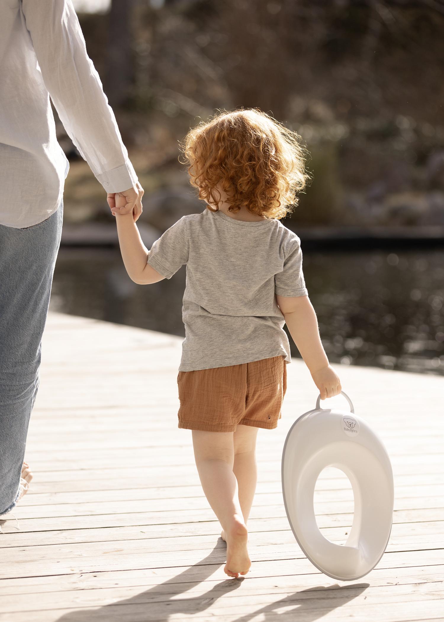 A child with curly red hair holds a white toilet seat and an adult's hand while walking on a wooden dock by the water.