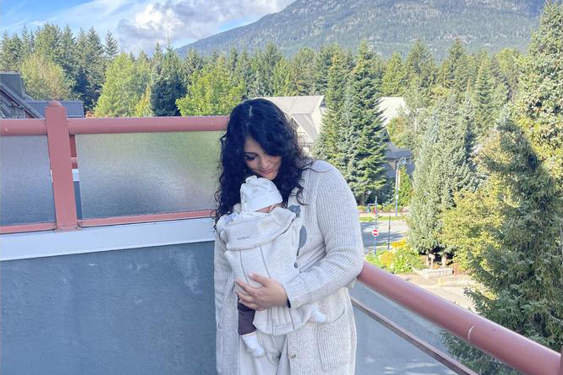 Woman in a light sweater cradles a baby in a front carrier on a balcony overlooking forested mountains.