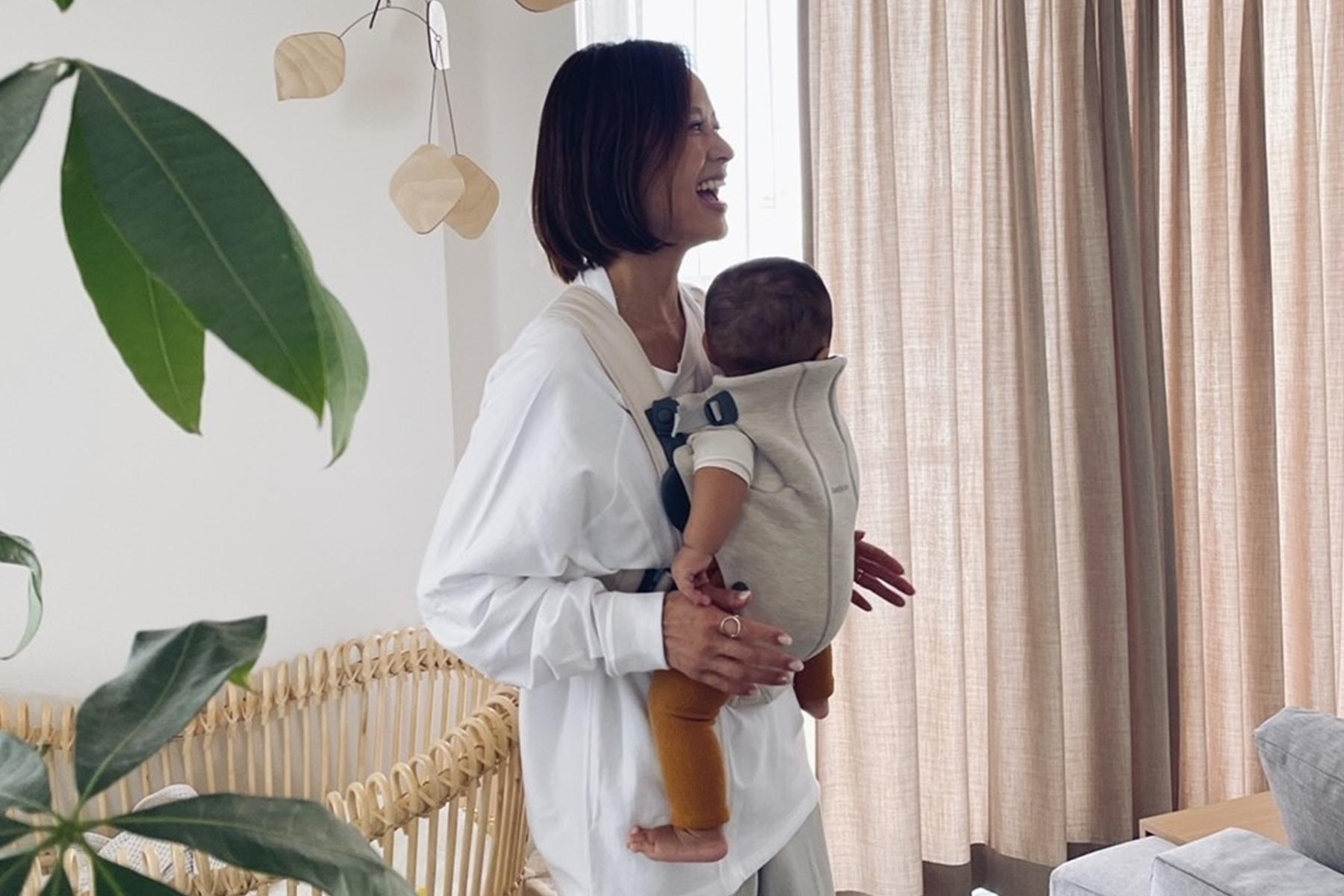 A woman in a white shirt smiles while holding a baby in a beige carrier. They are indoors near a window with beige curtains and plants.