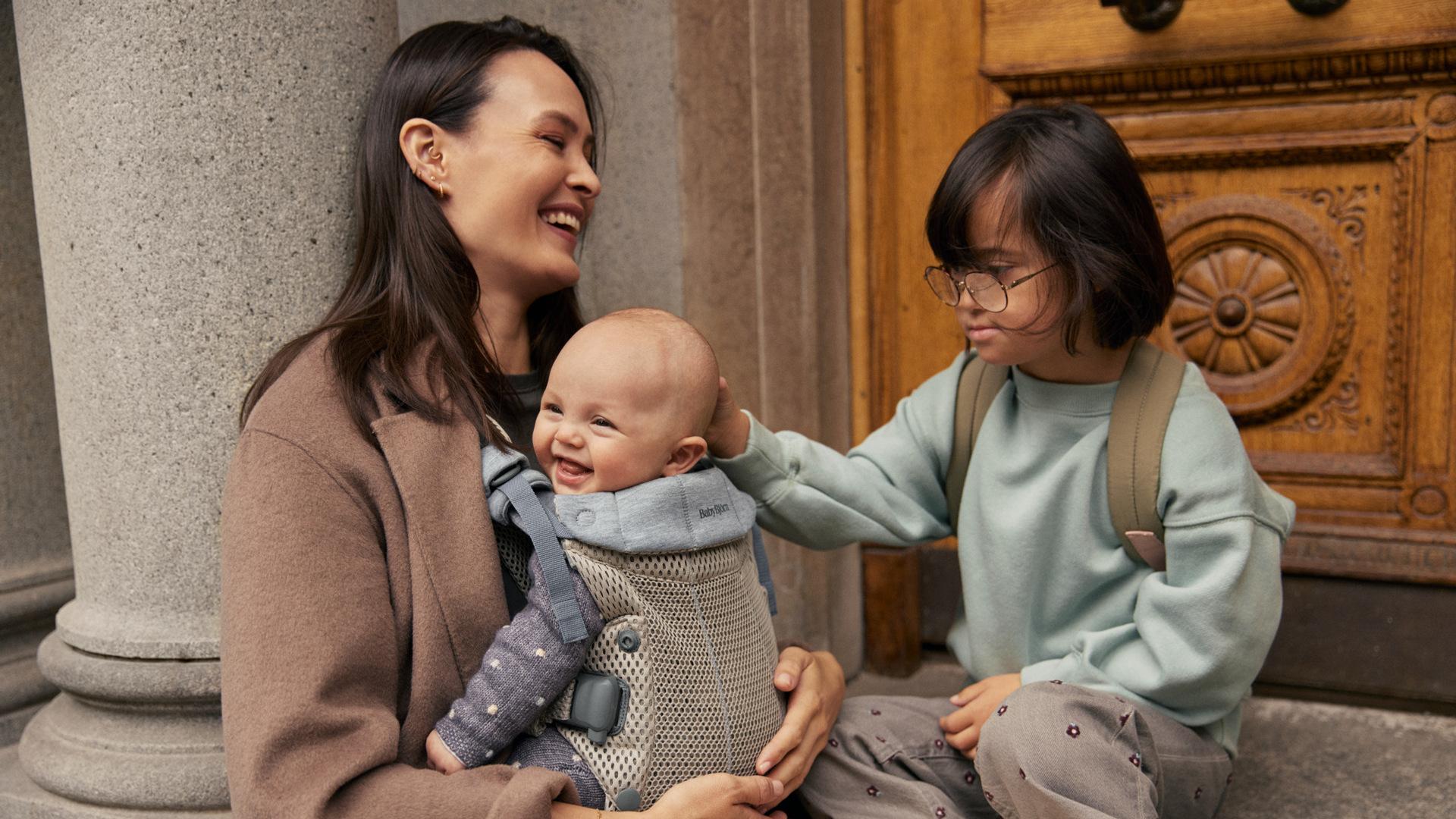 A woman sits on steps, holding a laughing baby in a carrier, while a child with glasses gently touches the baby's head.