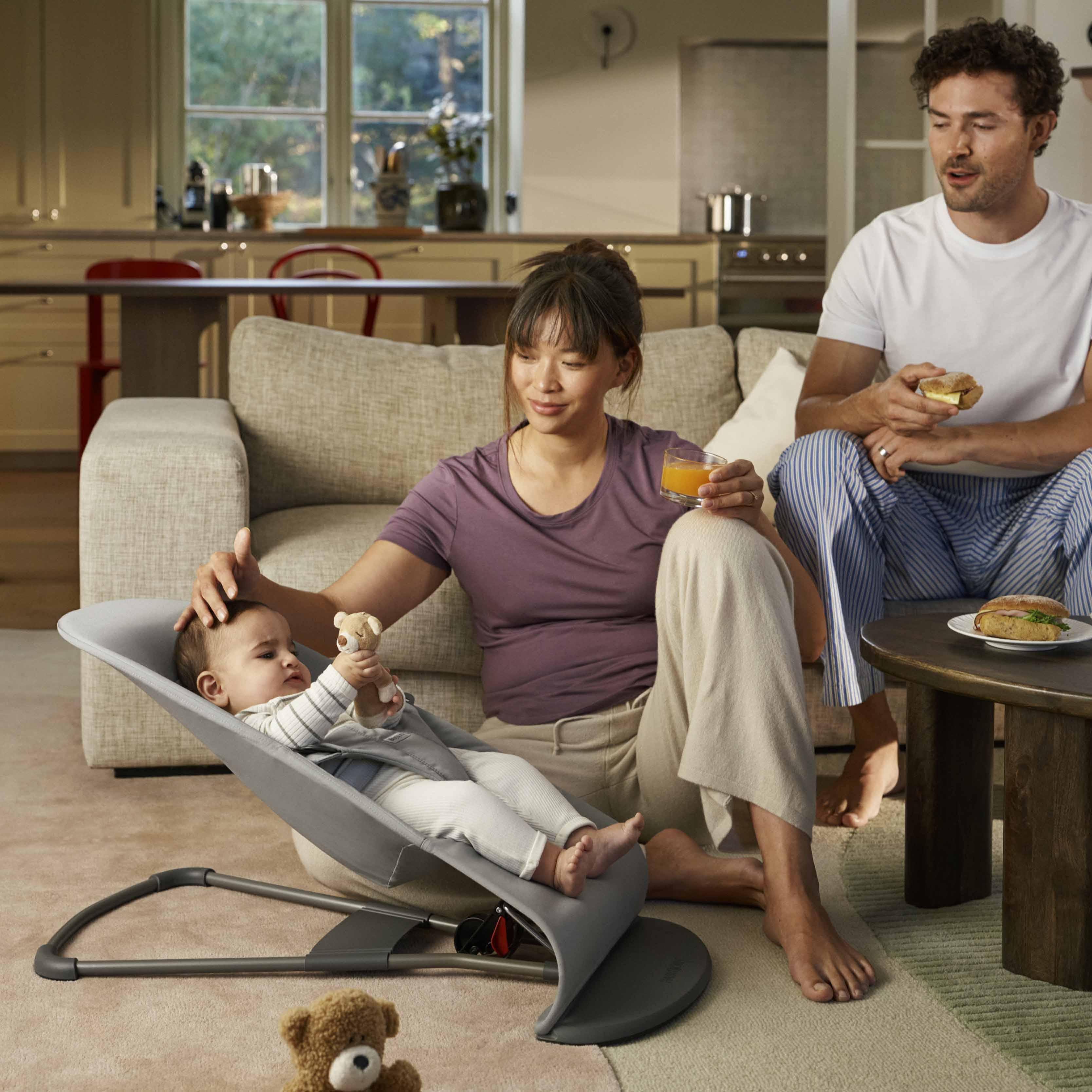 A woman and a man sit in a cozy living room, engaging with a baby in a bouncer. The woman holds a toy, while the man has a sandwich and an apple.